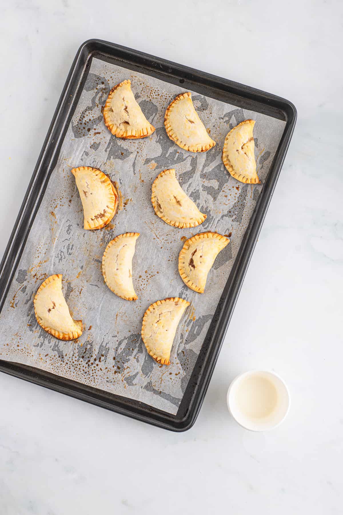 Baked pies on a sheet with glaze mixture sitting to the side.