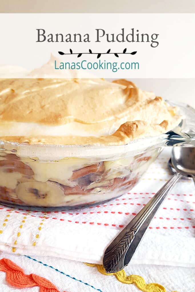 Banana pudding in a glass bowl on a kitchen towel with spoons in the foreground.