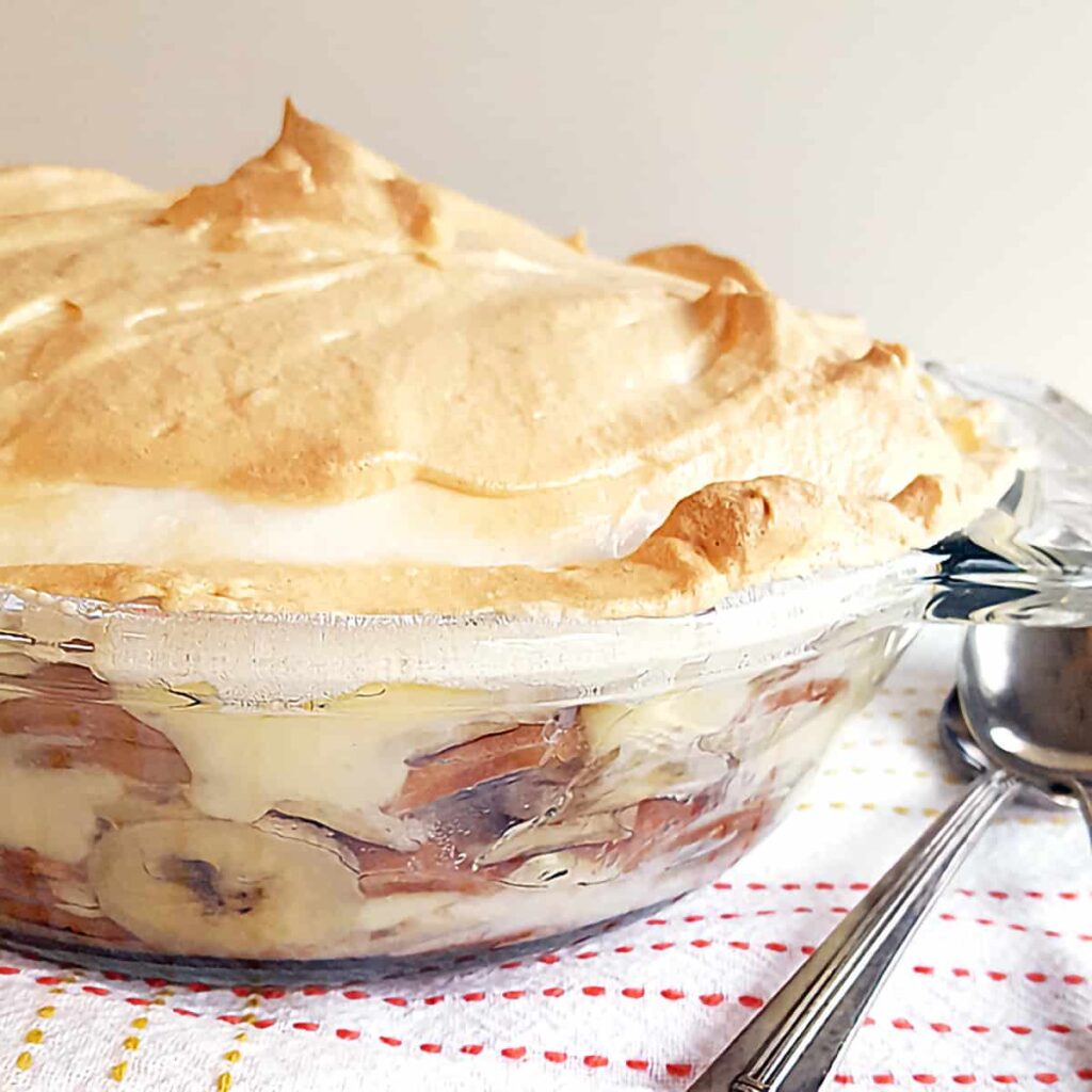 Banana pudding in a glass bowl on a kitchen towel with spoons in the foreground.