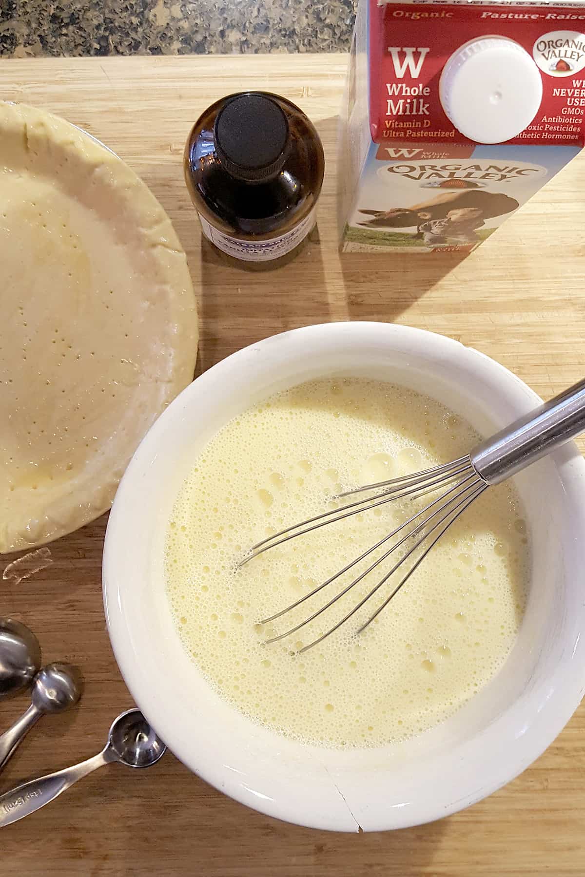 Medium mixing bowl holding the custard mixture. In the background are the prepared pie crust, a set of measuring spoons, vanilla extract, and milk.