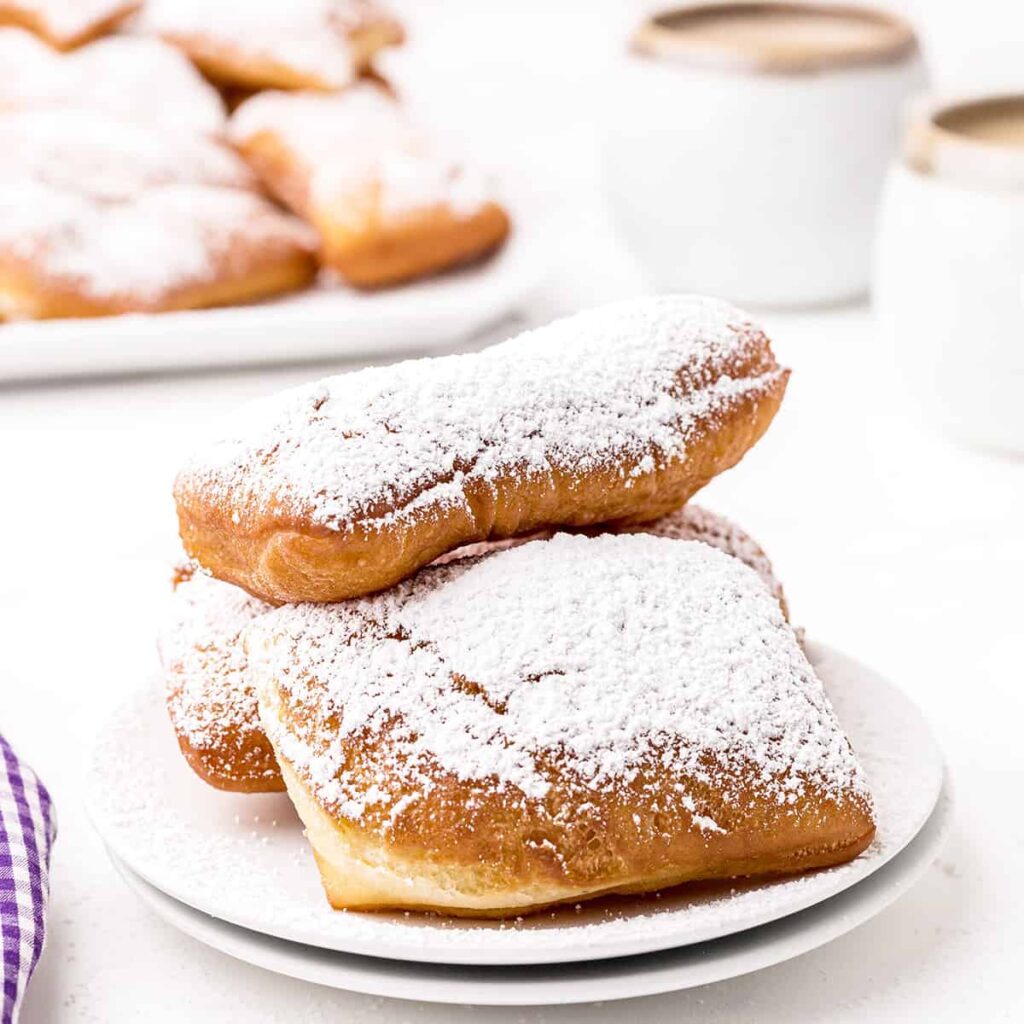 A serving of beignets topped with powdered sugar with a cafe au lait in the background.