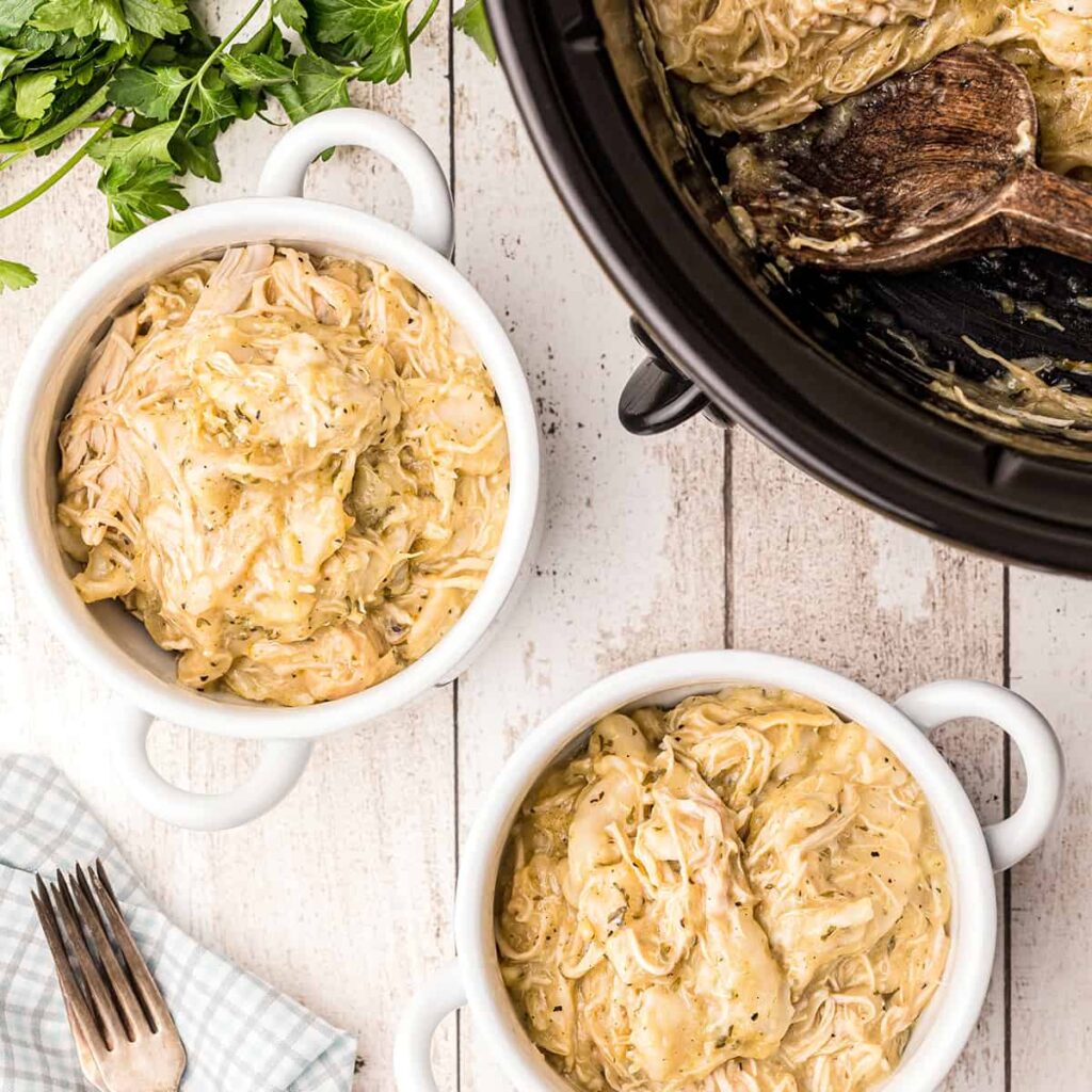 Two servings of chicken and dumplings in white bowls with a slow cooker in the background.